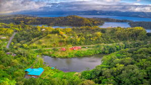 Aerial drone view of the Lucky Bug Property in Costa Rica, surrounded by lush tropical forest, with the bed and breakfast, gallery, and nearby lake nestled within the greenery.